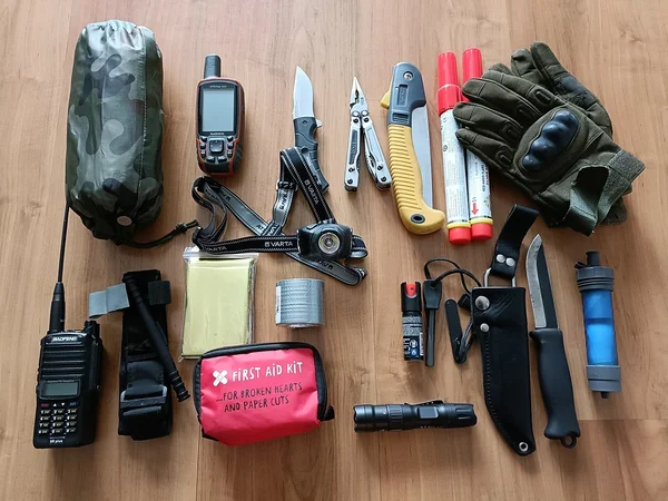 A survival gear set laid out on a wooden floor, including items like a GPS device, a multitool, a folding knife, gloves, a flashlight, a first aid kit, paracord, duct tape, a water filter, and a hand crank radio.