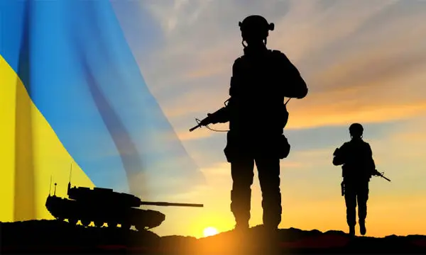 Silhouettes of two soldiers and a tank against the backdrop of a Ukrainian flag, with a dramatic sunset sky in the background.