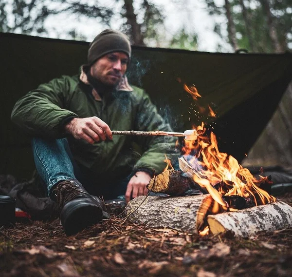 A man in outdoor gear is sitting in a forest, roasting a marshmallow over a campfire. Behind him is a makeshift shelter made of a tarp. The scene captures the essence of wilderness survival, with the man appearing focused and comfortable in his natural surroundings.